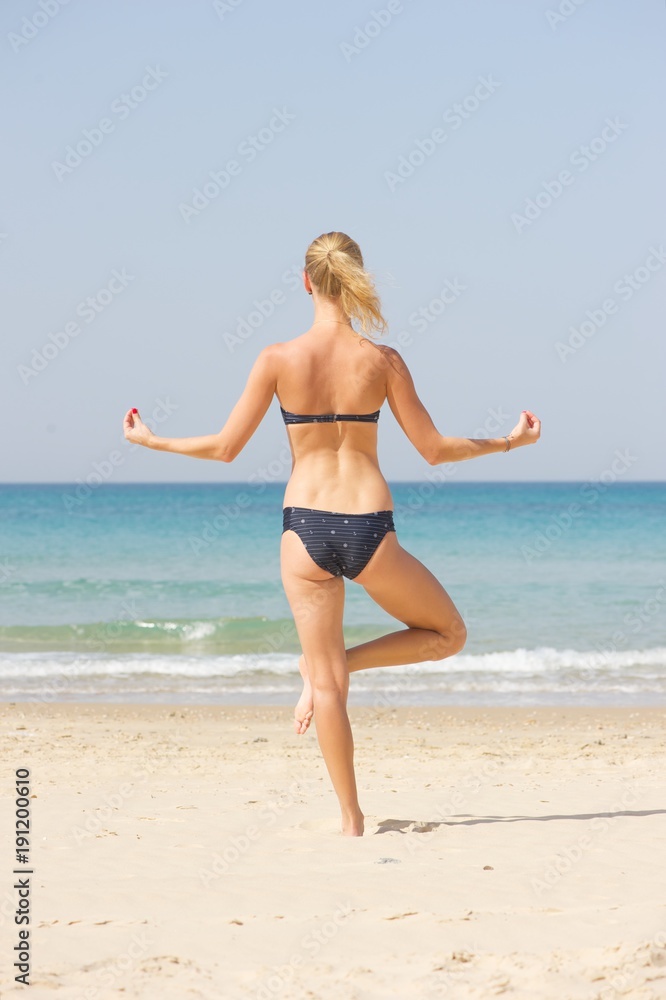 A young woman standing on one leg with her back to the camera; wearing  bikini; sea in background. Stock Photo | Adobe Stock