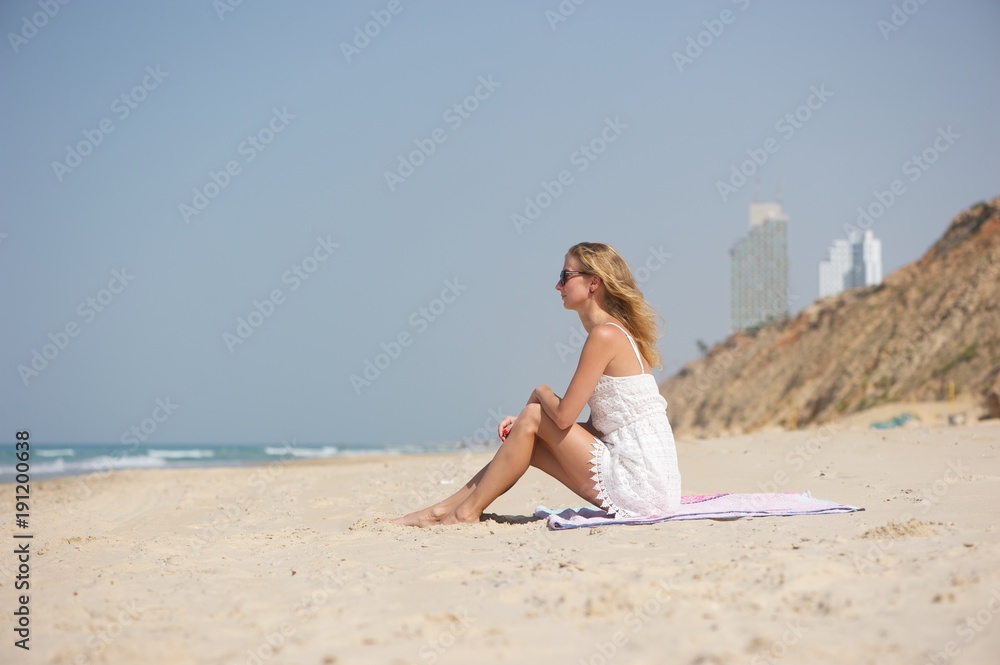A young woman wearing white lacy dress and sunglasses, sitting on a pink blanket at the beach; sea and hotels visible in background.
