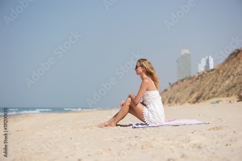 A young woman wearing white lacy dress and sunglasses  sitting on a pink blanket at the beach  sea and hotels visible in background.