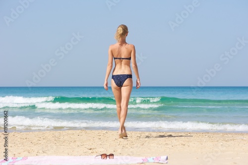 A day at the beach. A slim young woman in a biniki walking towards the sea. Pink blanket with sunglasses on it left behind her.