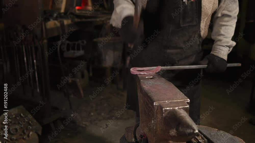 The blacksmith is froging red hot metal in a his forge with a big hammer. Hammering big metal hook on anvil. The blacksmith is froging red hot metal in a his forge with a big hammer. Hammering big metal hook on anvil.