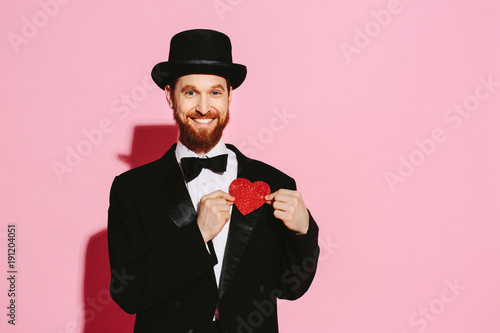 Smiling man in a tuxedo and top hat holding a red heart