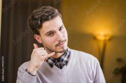 Attractive young man doing call me sign with hand and fingers, cheerful expression looking at camera