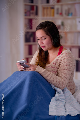 Close up of young beautiful woman with sleeplessness sitting on the bed using her cellphone, insomnia concept