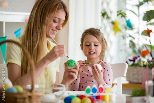 A mother and daughter celebrating Easter, painting eggs with brush. Happy family smiling and laughing. Cute little girl in bunny ears preparing the holiday.