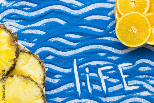 Life inscription on a blue colored sand with waves, slices of orange and pineapple, top view, flat lay photo