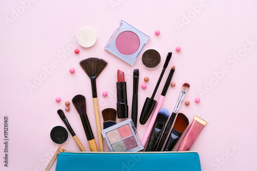 A blue cosmetics bag with makeup products spilling out on to a pastel pink background. Top view