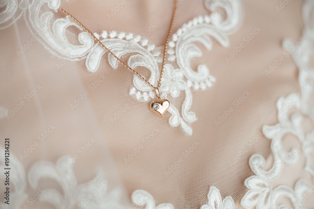Closeup view of cute small romantic golden heart hanging on necklace chain on breast of young bride. Woman wears elegant white wedding dress. Horizontal color image.