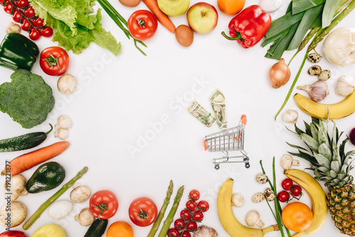 top view of small shopping cart with dollars between vegetables and fruits isolated on white