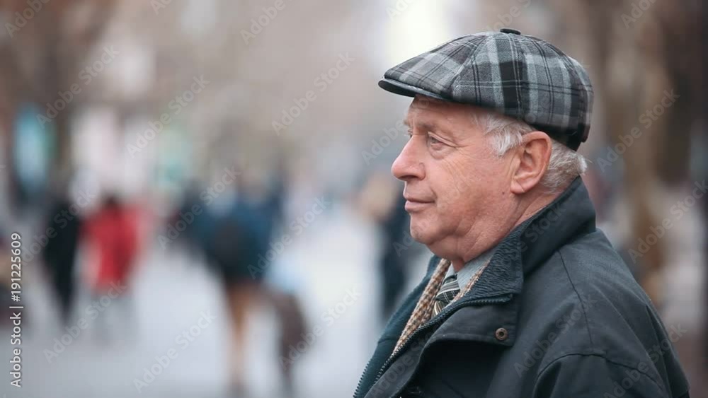 A profile view of a senior man dressed in a black zipup coat and a woollen flat cap. He stands on a park alley with people in autumn. A profile view of a senior man dressed in a black zipup coat and a woollen flat cap. He stands on a park alley with people in autumn.