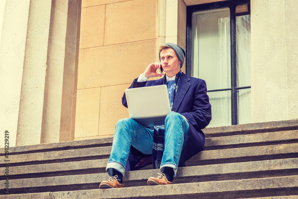 American man traveling, working in New York, wearing blue long woolen overcoat, scarf, jeans, leather shoes, knit hat, sits on stairs outside on street, works on laptop computer, talks on cell phone..