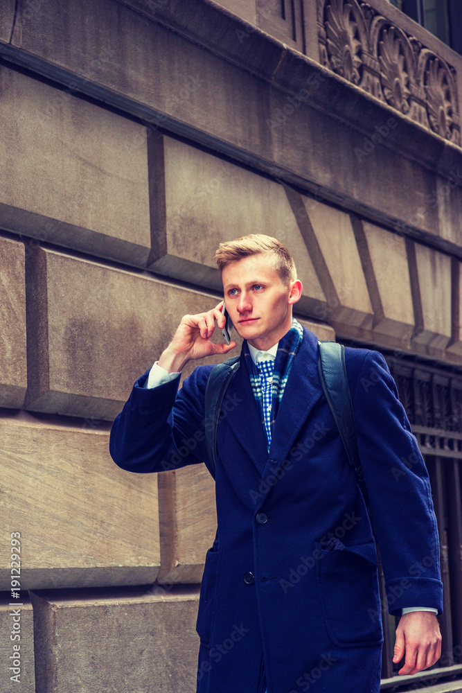 Young American college student traveling in New York, wearing blue long woolen overcoat, scarf, carrying back bag, walking on vintage street in winter, talking on cell phone. .