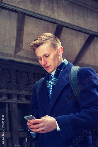 American college student traveling in New York, wearing blue long woolen overcoat, scarf, reading messages on cell phone while walking on street in winter..