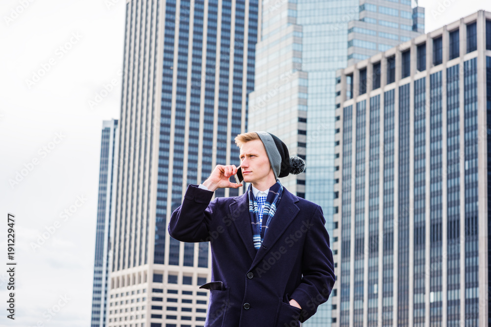 Young American man traveling in New York, wearing blue long woolen overcoat, scarf, cuffed knit beanie hat with pom pom, standing in front of business district, talking on cell phone. . Young American man traveling in New York, wearing blue long woolen overcoat, scarf, cuffed knit beanie hat with pom pom, standing in front of business district, talking on cell phone. .