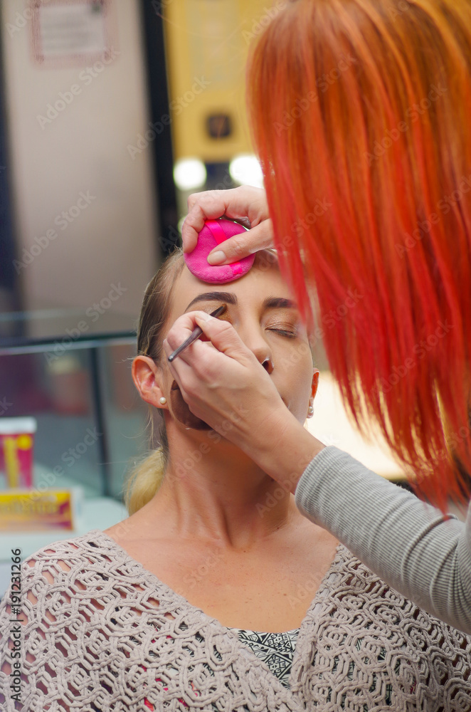 Fototapeta premium Close up of beautiful face of young woman getting make-up. The artist is applying eye mascara on her eyes. The lady closed eyes with relaxation