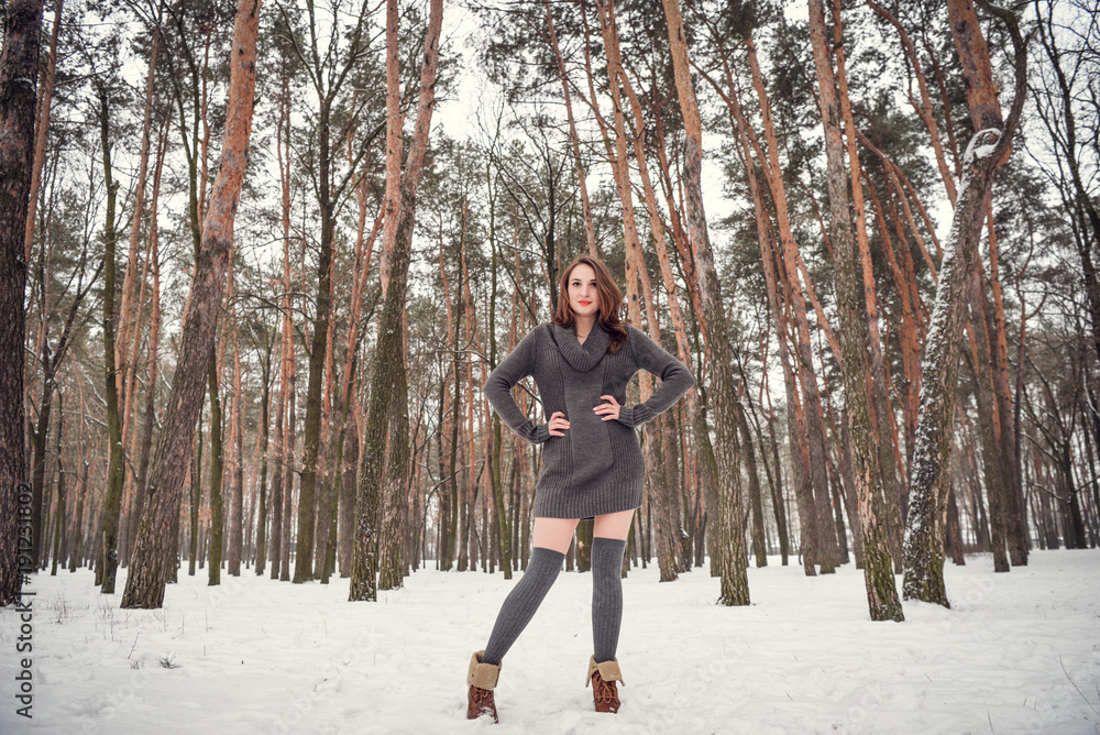 Winter portrait of a happy beautiful girl with brown hair in snow winter forest . Woman wearing knitted grey pullover dress and stockings high socks. Winter atmosphere, nature and freedom concept.