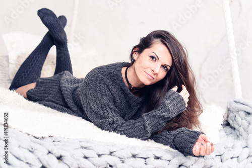 Beautiful sexy tan brunette woman posing in bedroom, wearing cozy grey sweater and knitted socks. Rest, sleeping, comfort concept. Close up of young woman lying in bed at home. Scandinavian home. 