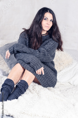 Beautiful sexy tan brunette woman posing in bedroom, wearing cozy grey sweater and knitted socks. Rest, sleeping, comfort concept. Close up of young woman lying in bed at home. Scandinavian home. 
