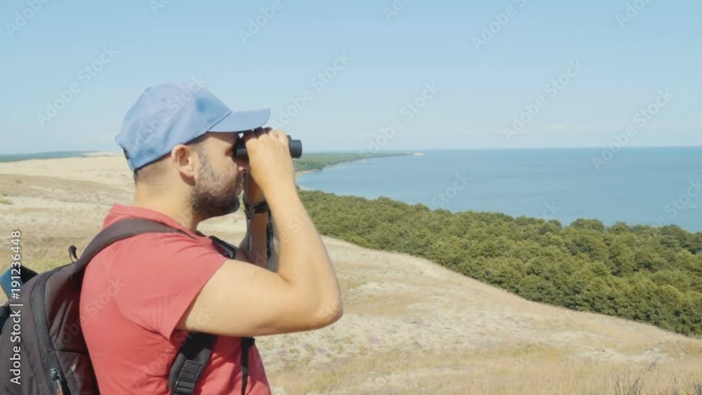 Travel man looking at wildlife with binoculars. Male on vacation looking at nature landscape. Hiker with a backpack is looking through binoculars at sea, at beautiful landscape. Mountain. Beach. Travel man looking at wildlife with binoculars. Male on vacation looking at nature landscape. Hiker with a backpack is looking through binoculars at sea, at beautiful landscape. Mountain. Beach.
