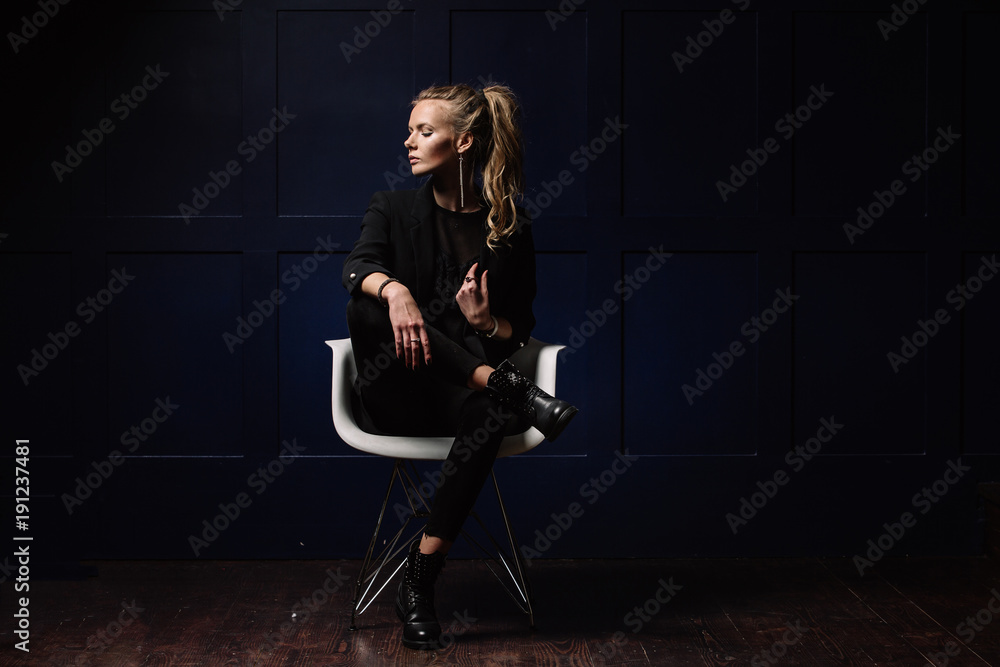 Sexy young model in dark clothes, sitting on a white chair and posing on a dark blue background. Watch on hand. Fashion, Beauty, Style Sexy young model in dark clothes, sitting on a white chair and posing on a dark blue background. Watch on hand. Fashion, Beauty, Style