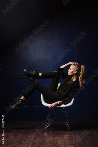 Sexy young model in dark clothes, sitting on a white chair and posing on a dark blue background. Watch on hand. Fashion, Beauty, Style