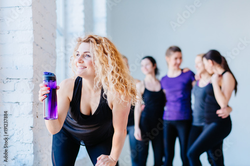 Attractive happy blond curly woman drinking water from the bottle in gym. Teamwork, good mood and healthy lifestyle concept. © Iryna