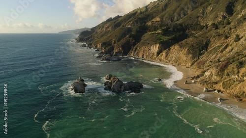 Aerial shot flying down towards sandy beach with rock arches coming out of the ocean. Hilly coastline, beautiful day.