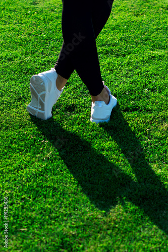 Sports Legs of young athletic woman in dark tights and light sneakers stay or walking along path among dense green grass in training. Copy space. Healthy lifestyle concept. European female