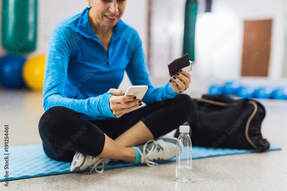 Mature woman taking a break and eating chocolate in gym Mature woman taking a break and eating chocolate in gym