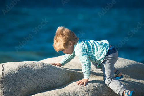 Child climbing stone. Cute fashionable baby boy in blue shirt and gray pants climbs on stone at sunny summer day on natural sea background. Young blond boy have fun on seaside. Sport for children