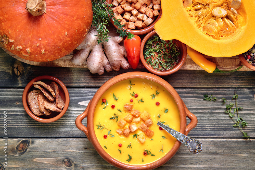 Pumpkin soup in ceramic bowl on rustic wood served with thyme and croutons. Next to it is a tray with healthy soup ingredients. Flat lay, view from above. Pumpkin soup in ceramic bowl on rustic wood served with thyme and croutons. Next to it is a tray with healthy soup ingredients. Flat lay, view from above.