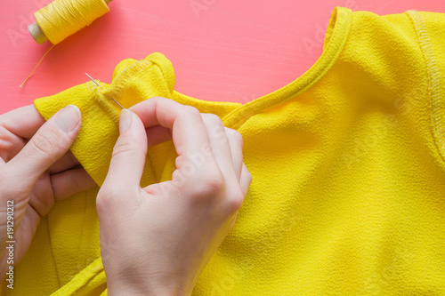 Hand with a needle and thread sewing a yellow blouse on the pastel pink table in an atelier. Handmade work. Womanly hobby. Professional sewing concept.