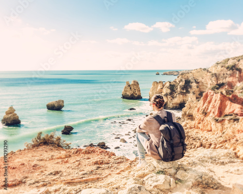 Girl traveler with a backpack sits on the rocks on the ocean, admiring the incredible scenery. Portugal, the Algarve, a popular destination for travel in Europe
