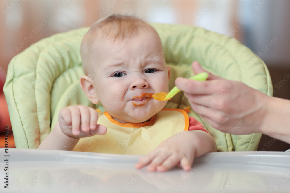 Cute baby boy refusing to eat food from spoon with face dirty of vegetable puree.