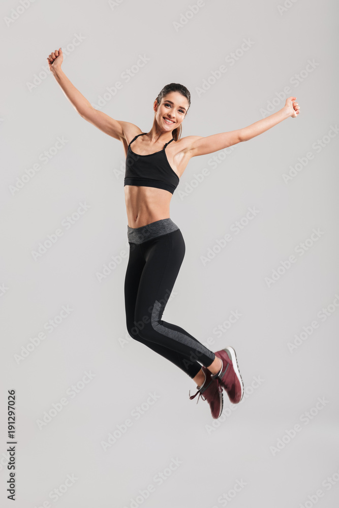 Full-length photo of happy healthy woman in sportswear with abs jumping and having fun in gym, isolated against gray background