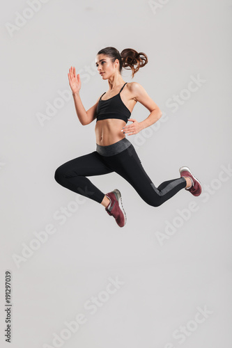 Fototapeta Naklejka Na Ścianę i Meble -  Full-length photo in profile of happy healthy woman in sportswear with abs jumping and flying in studio, isolated over gray background