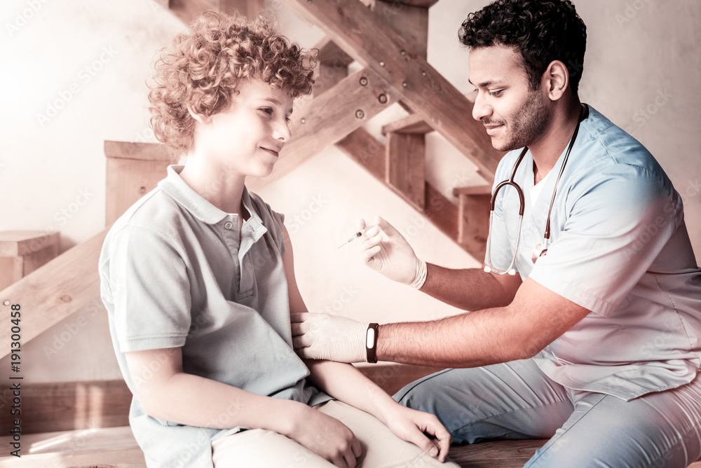 Fototapeta premium Do not worry. Cheerful doctor smiling while getting ready to vaccinate a curly haired boy indoors.