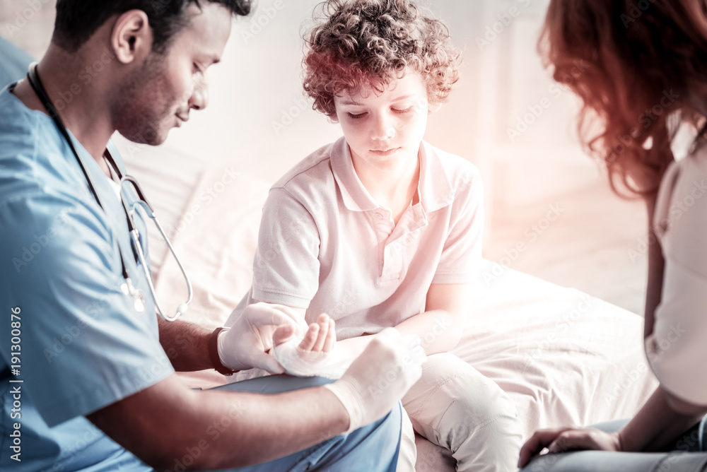 Traumatic moment. Selective focus on a sad curly haired boy looking at a doctor implying a bandage on his arm Traumatic moment. Selective focus on a sad curly haired boy looking at a doctor implying a bandage on his arm