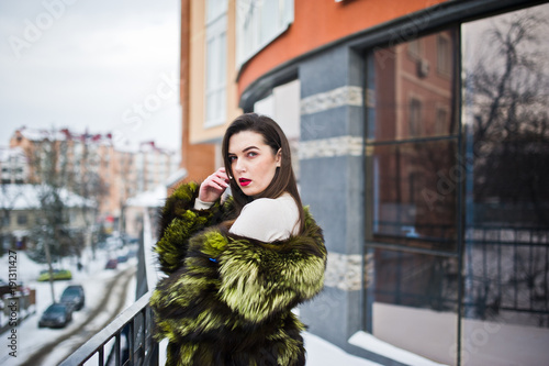 Brunette girl in green fur coat at street of city against house with large windows at winter. © AS Photo Family