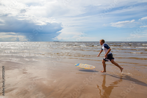 Young sportive man is running after his yellow and blue skimboard on the beach on a sunny day and on the sky there are dark clouds photo