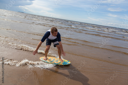 Young sportive man is sliding on yellow blue skimboard on the baltic sea beach and there are dark clouds on the sky but sunny