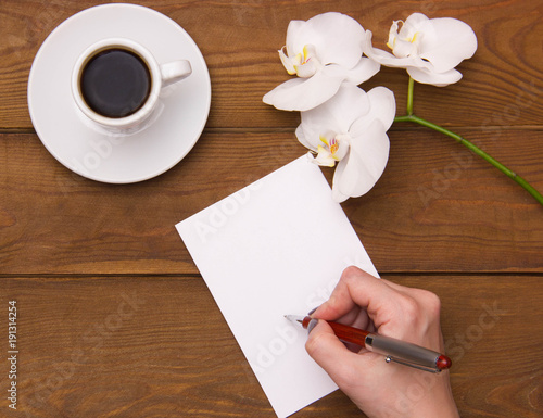 female hand writing a note in a clean white notepad pen on a wooden table next to a cup of coffee and white orchid flowers