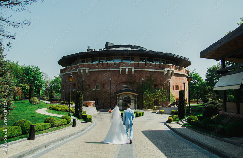 Wedding couple is walking holding hands near the old ancient citadel castle on sunny day. Bride in puffy dress and groom in light blue suit. Picturesque architecture and view. Green lawn.