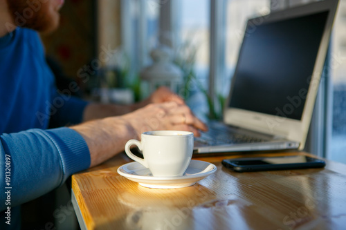 Close-up of men's hands, typing on a laptop, drinking coffee, next to the phone. A cup of coffee is empty. Close-up. Sunlight