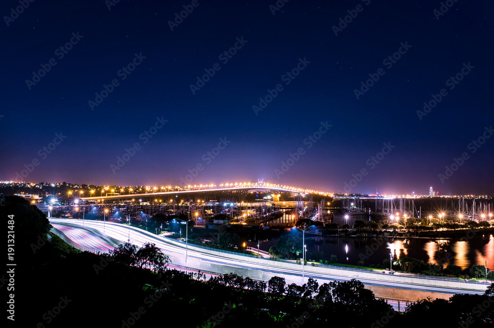 cars in motion on a lighted bridge and highway over the sea with anchored sailing boats in a marina of Auckland in New Zealand with a Starry sky