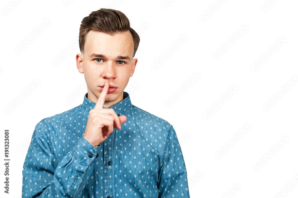 White young guy dressed in blue shirt showing silence gesture with his forefinger by mouth on a white background. Isolated. White young guy dressed in blue shirt showing silence gesture with his forefinger by mouth on a white background. Isolated.