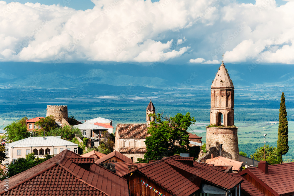 Fototapeta premium Old houses with brown tile roofs, church of St. George in city of love - Sighnaghi, Kakheti, Georgia. Beautiful view on Alazani valley. Sunny day with blue sky with clouds.