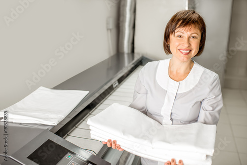 Portrait of a senior woman chambermaid standing with clean bedclothes near the professional ironing machine in the laundry
