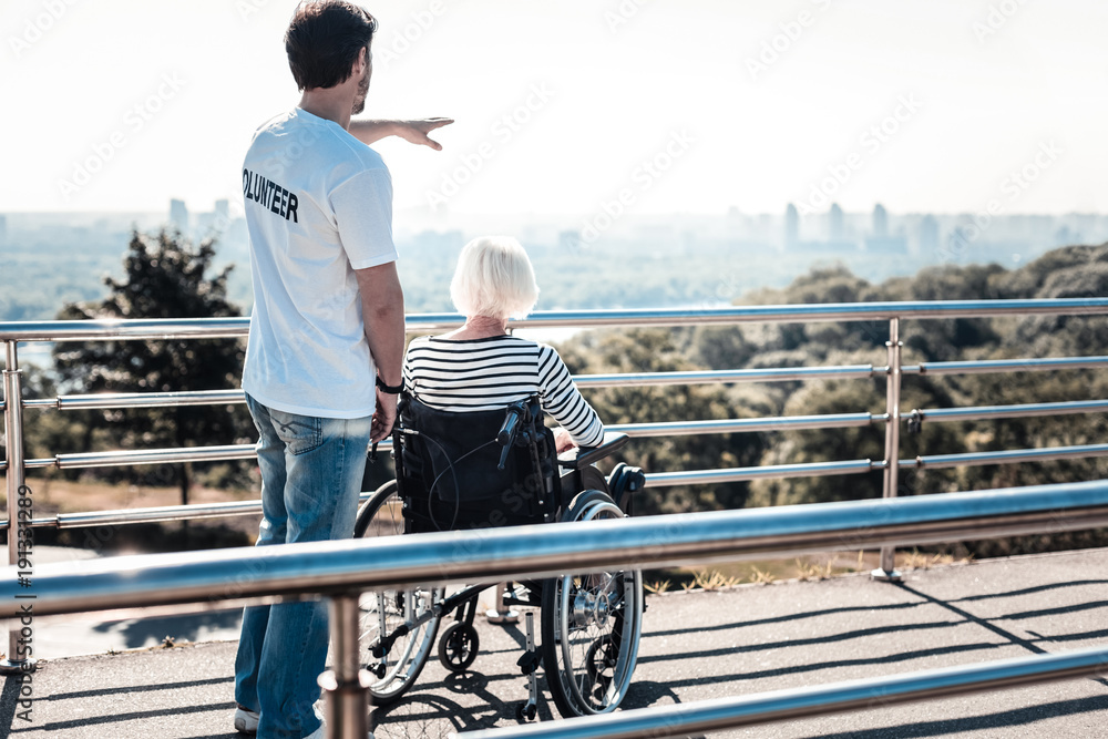 How beautiful. Nice positive handsome man looking at the view and pointing with his hand while having a walk with a disabled elderly woman How beautiful. Nice positive handsome man looking at the view and pointing with his hand while having a walk with a disabled elderly woman