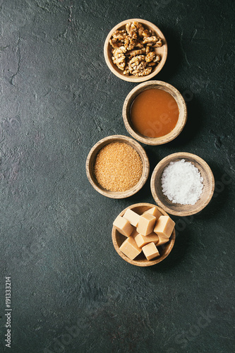 Salted caramel fudge candy served with fleur de sel, caramel sauce, brown cane sugar and caramelized walnuts in wood bowls over black texture background. Top view, space. Dessert set